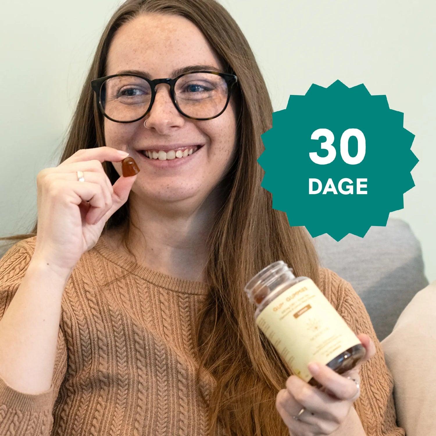 Kvinde med briller smiler og holder en kapsel og en flaske kosttilskud foran en 30-dages badge.