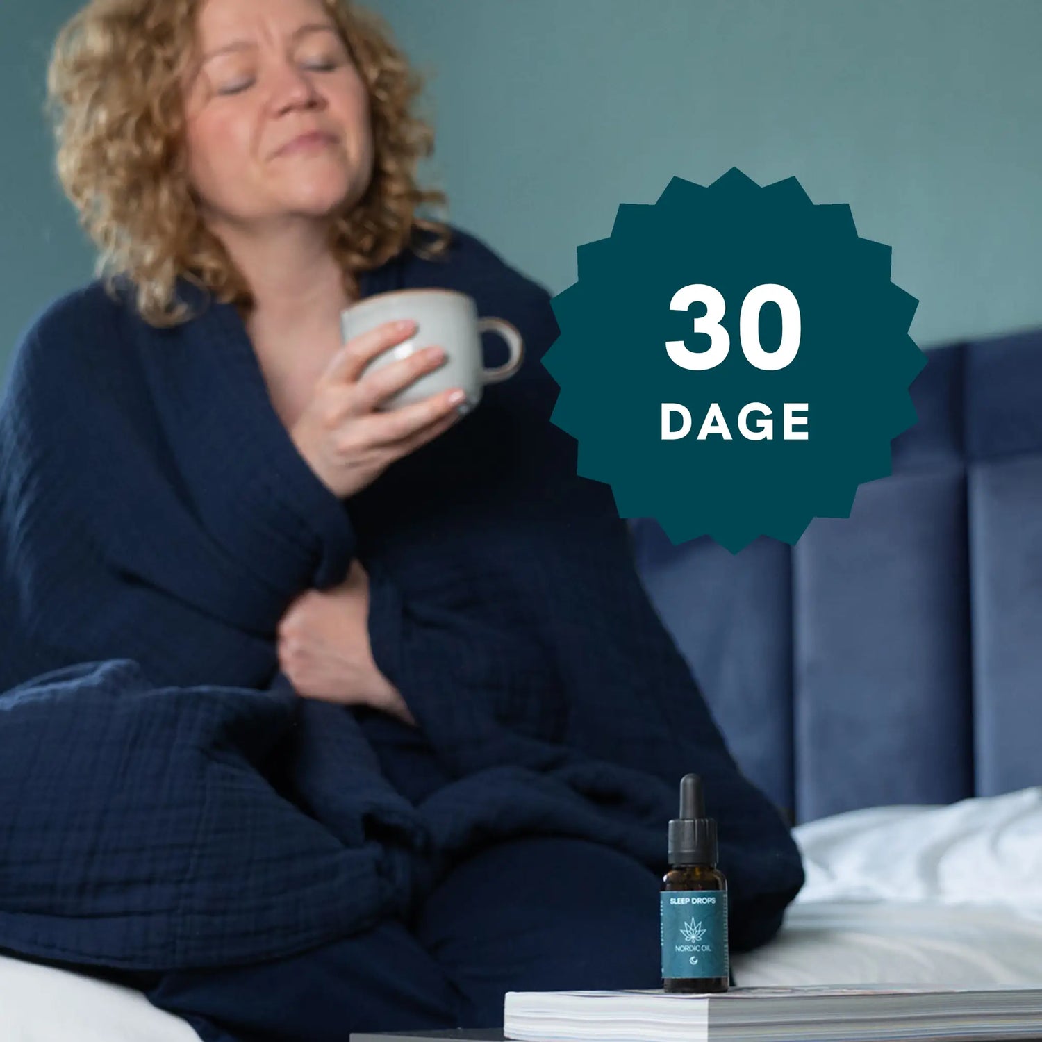 Kvinde i mørkeblå morgenkåbe sidder i sengen og holder en kop, med en Nordic Oil Sleep Drops-flaske og en 30-dages badge i nærheden.