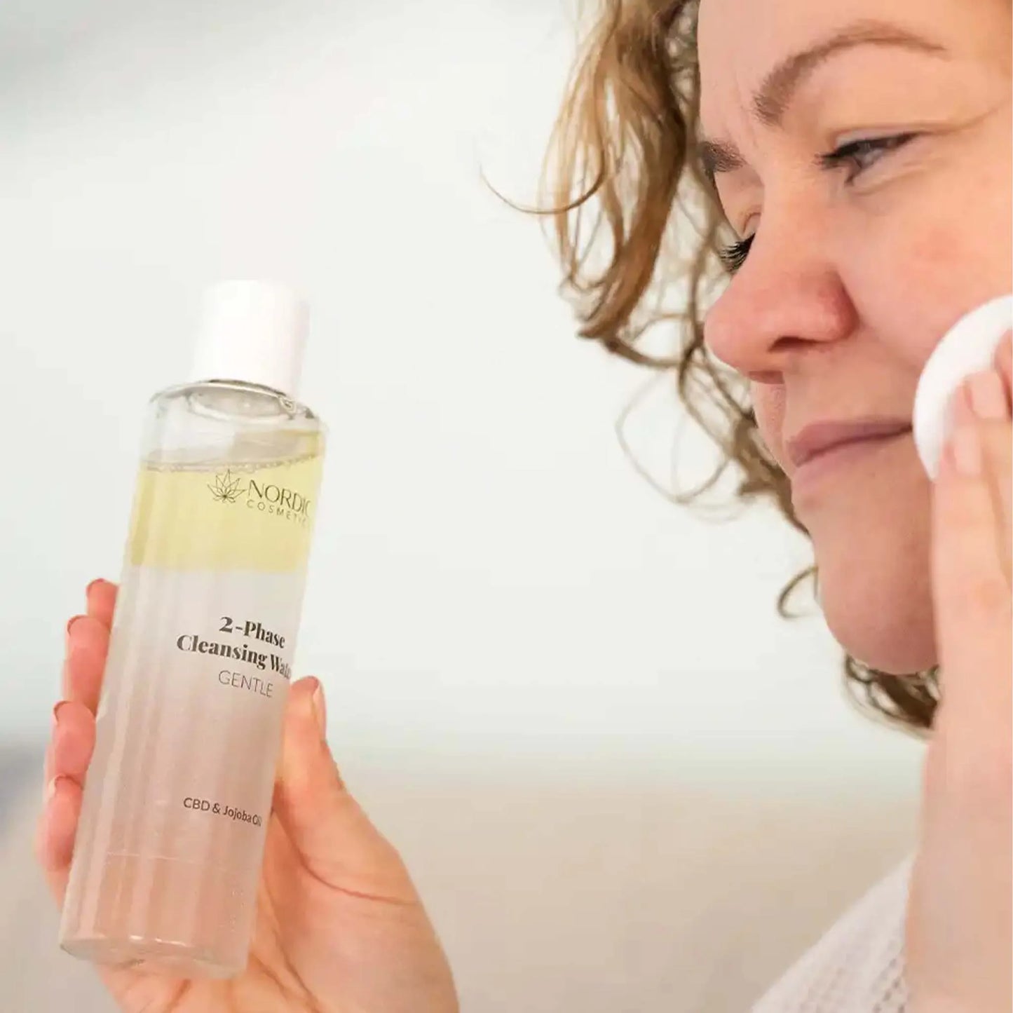 Kvinde der renser ansigtet med en bomuldspude ved siden af en flaske Nordic Cosmetics 2-Phase Cleansing Wash.