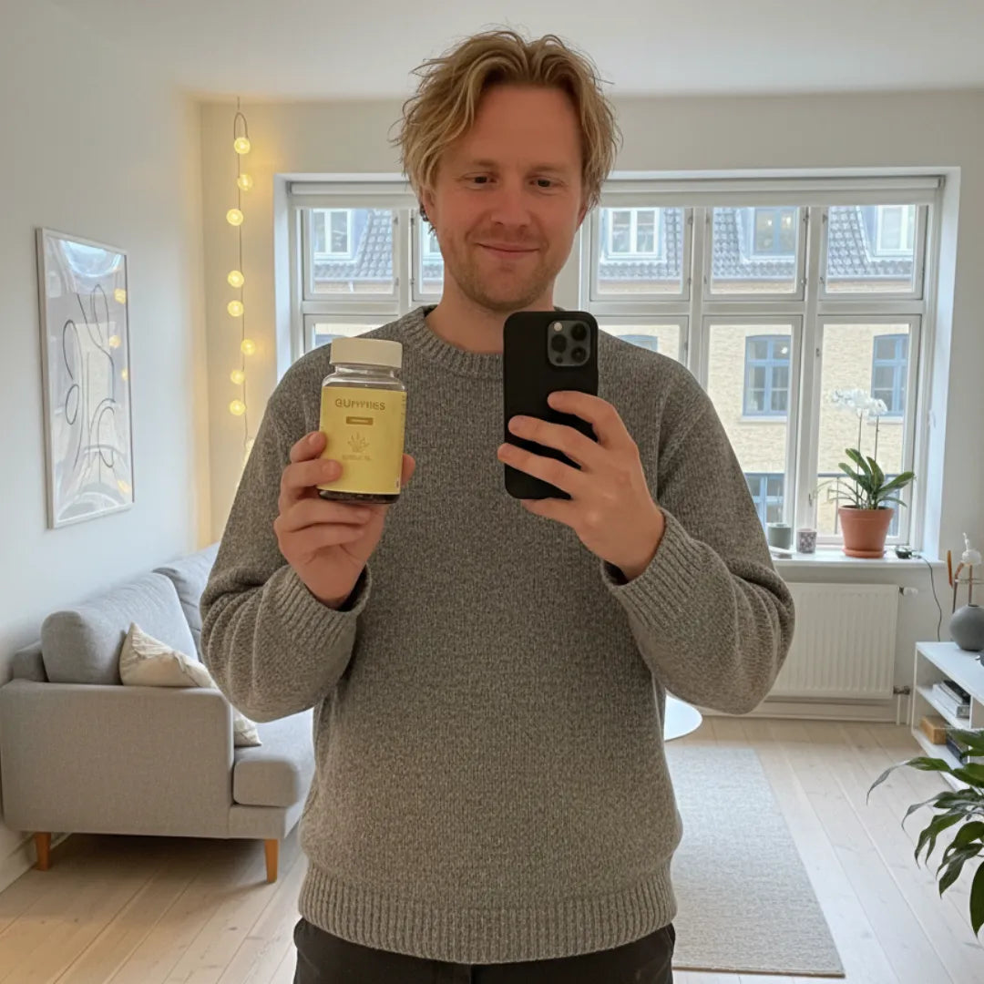 Mand i grå sweater tager en selfie i en lys stue og holder en flaske CBD-gummier.