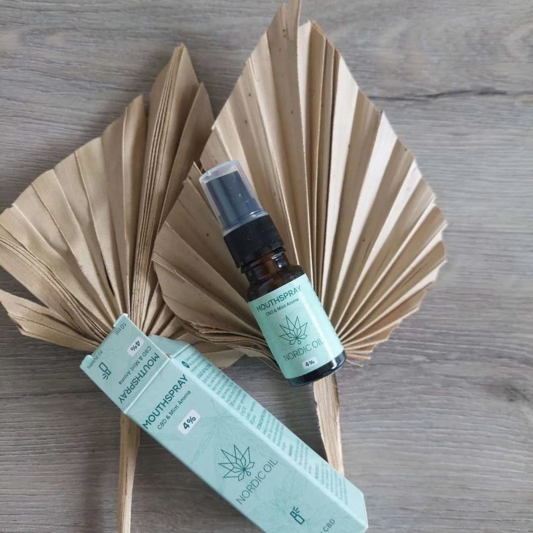 Mintfarvet Nordic Oil CBD mundspray med tilhørende æske på træbord, med tørrede palmeblade i baggrunden.