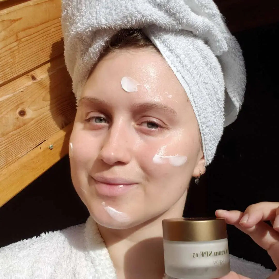 Kvinde med ansigtsmaske og håndklæde om hovedet, holder en krukke ansigtscreme.