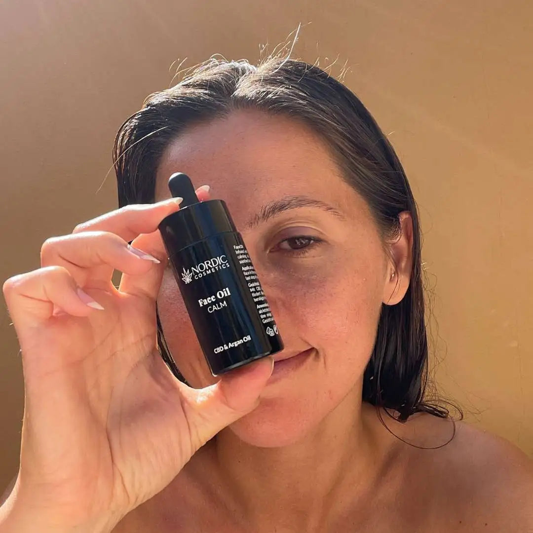 Kvinde holder en mørk flaske Nordic Cosmetics Face Oil Calm (CBD & Argan Oil) foran ansigtet.