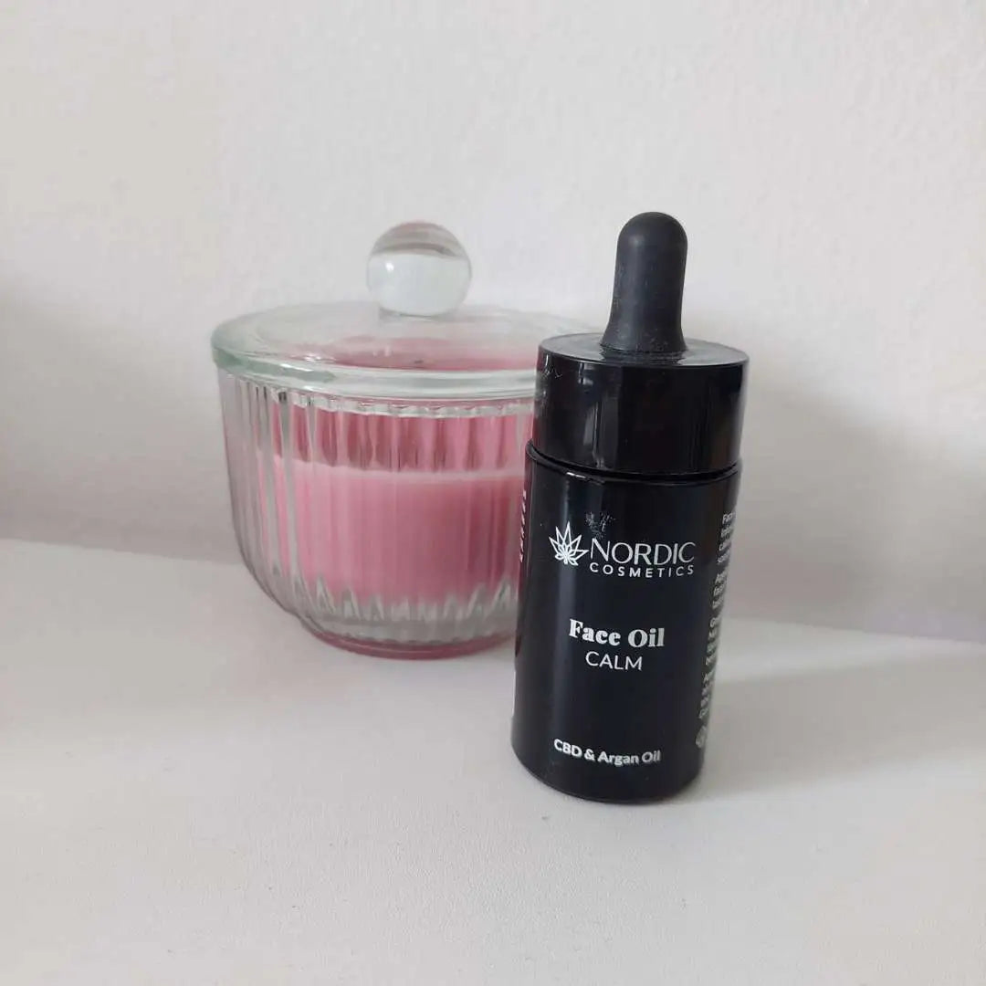 Sort dropperflaske med Nordic Cosmetics Face Oil Calm CBD og Argan Oil ved siden af en pink glasbeholder med låg.