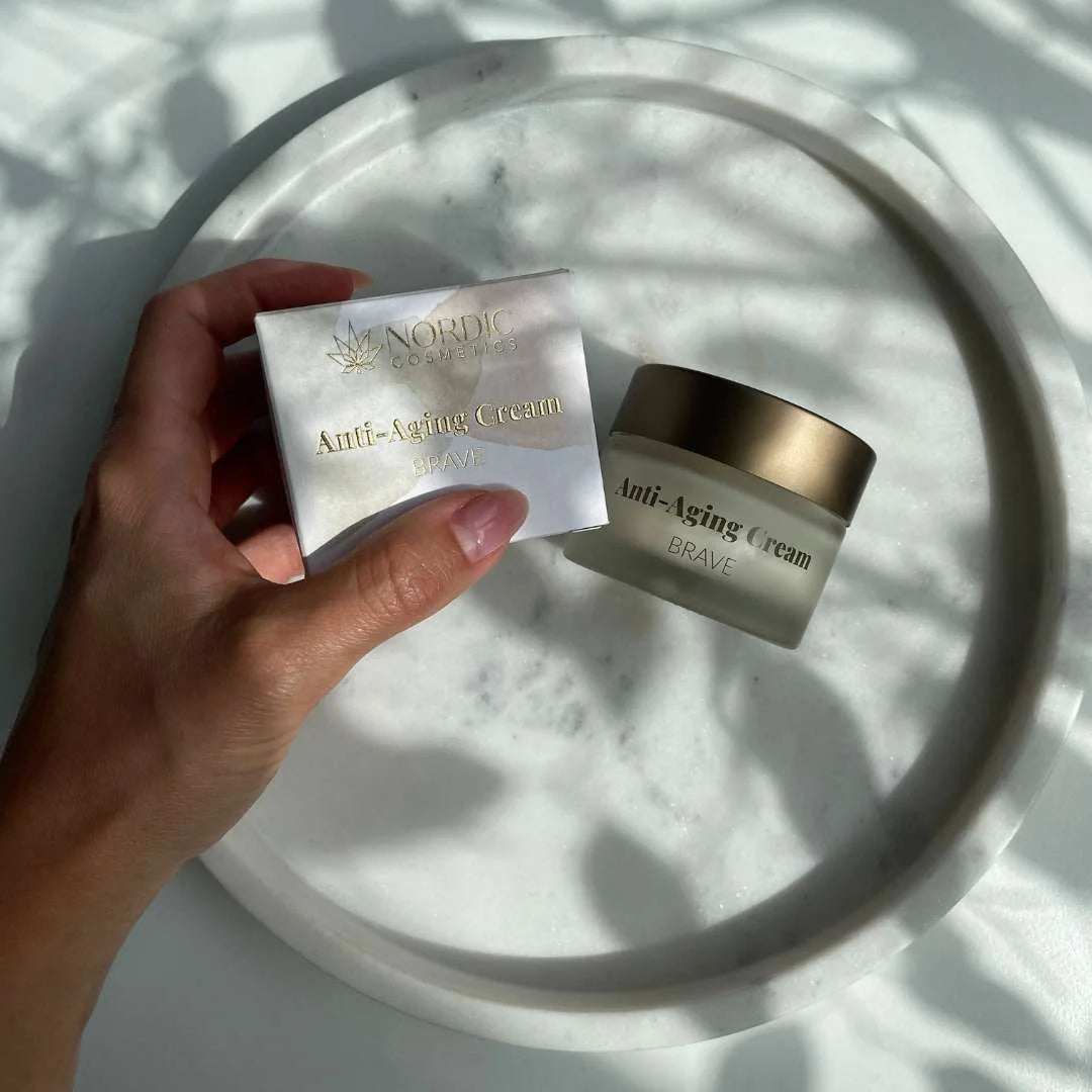 Hånd holder boks og glaskrukke med Nordic Cosmetics Anti-Aging Cream Brave på hvid marmorbakke