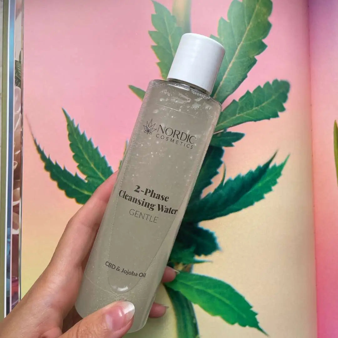 Nordic Cosmetics 2-Phase Cleansing Water – mildt rensevand med CBD og jojobaolie i gennemsigtig flaske, med cannabisblade i baggrunden.