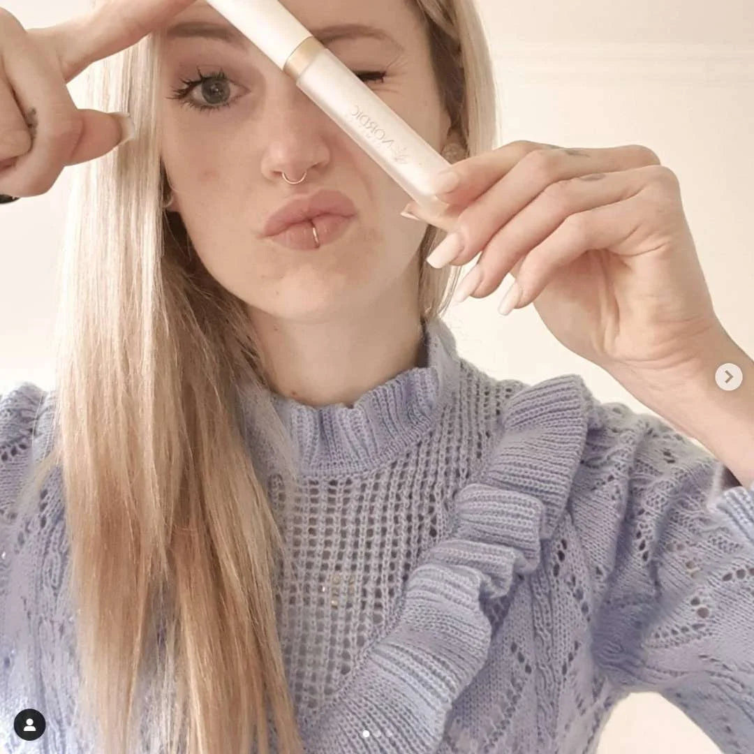 Kvinde med langt lyst hår holder en lys kosmetisk tube tæt ved ansigtet og laver et kysseudtryk.