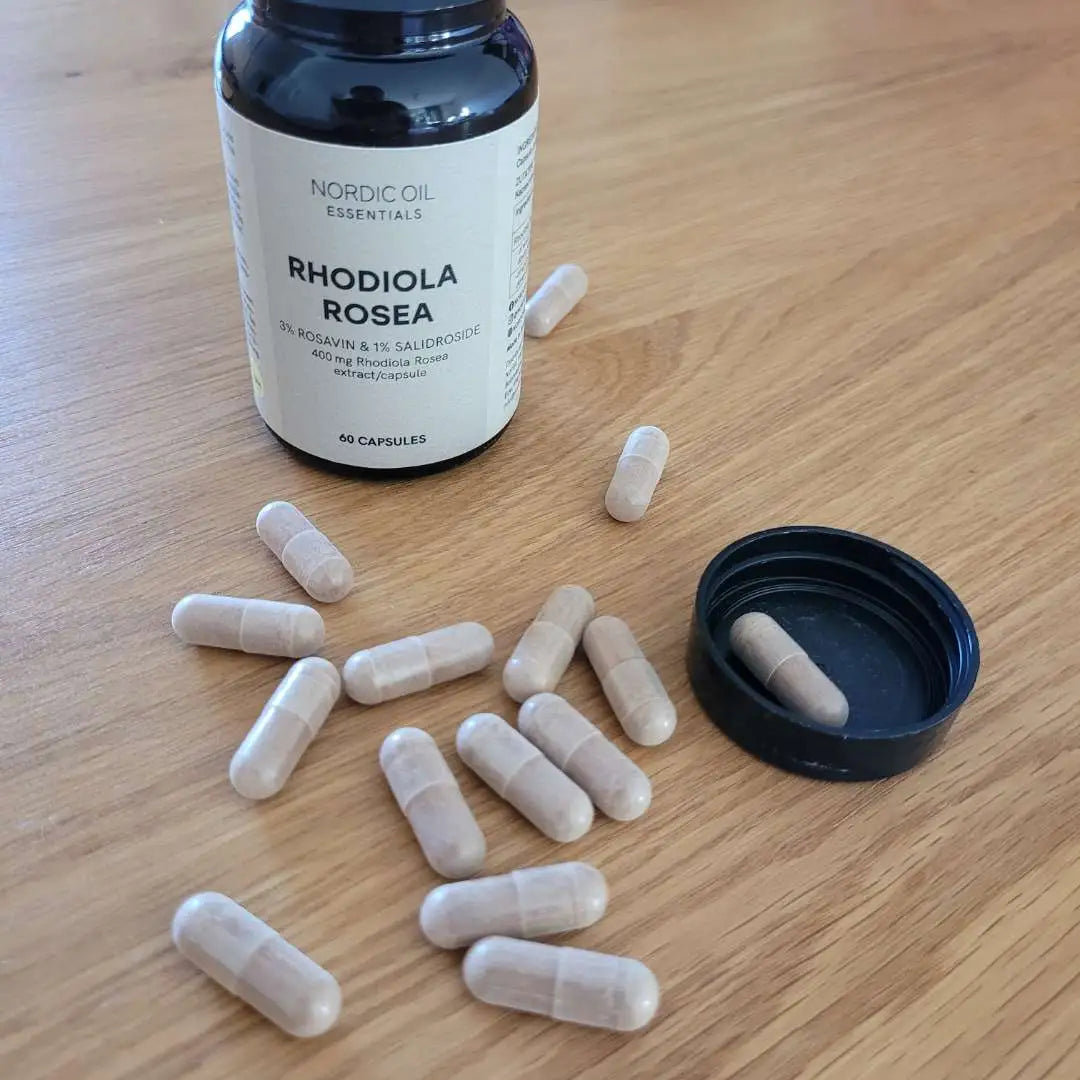Rhodiola rosea-kapsler spredt ud på et træbord ved siden af en blå flaske.