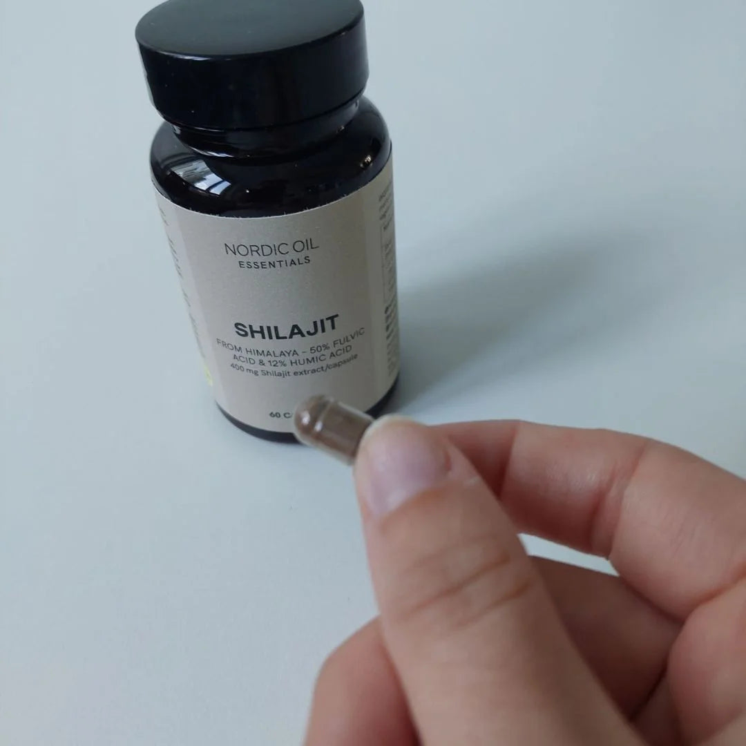 Shilajit-kapsel holdes mellem fingrene foran en Nordic Oil Essentials Shilajit-flaske.