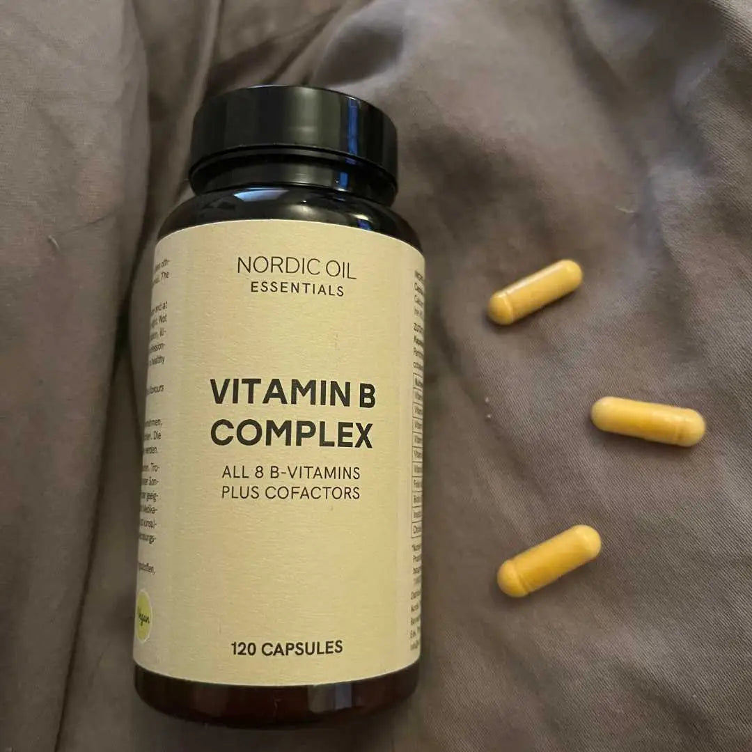 Nordic Oil Essentials Vitamin B-kompleks flaske med 120 kapsler og tre gule kapsler ved siden af på gråt stof