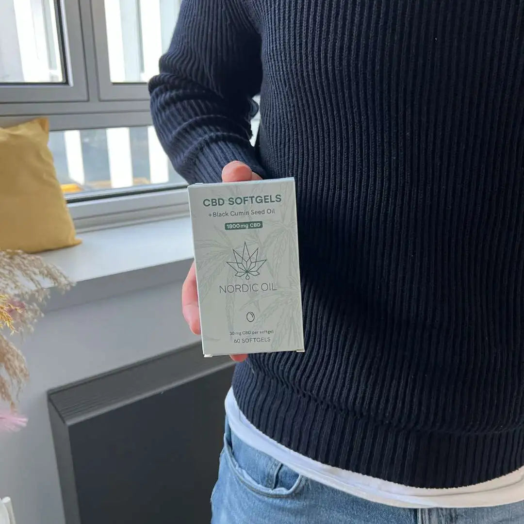 Person i en mørk trøje holder en Nordic Oil CBD-softgels-boks med sort kommen frøolie.