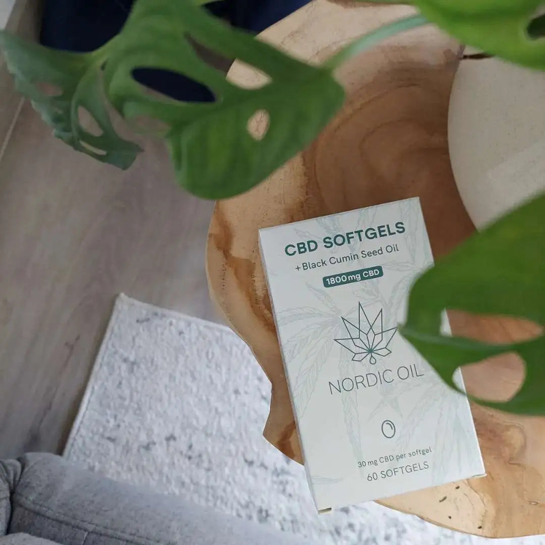 Nordic Oil CBD-softgelboks med sortkommenolie på træoverflade, omgivet af grønne blade