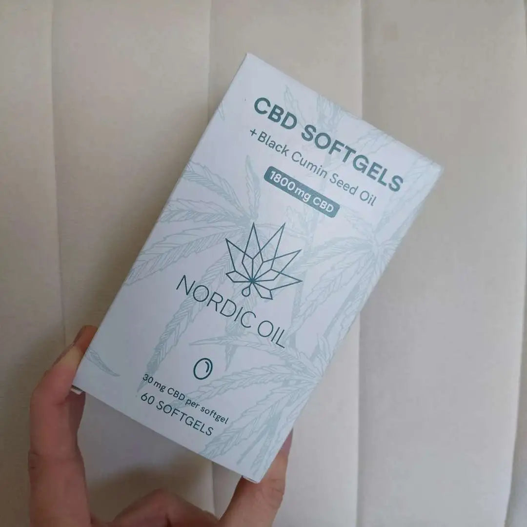Hvid boks med Nordic Oil CBD-softgels og sort kommenfrøolie, holdt i en hånd.