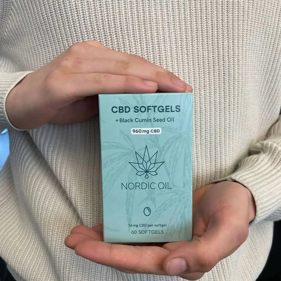 Lyseblå Nordic Oil CBD-softgels-æske med sort kommenolie, holdt af person i beige trøje.