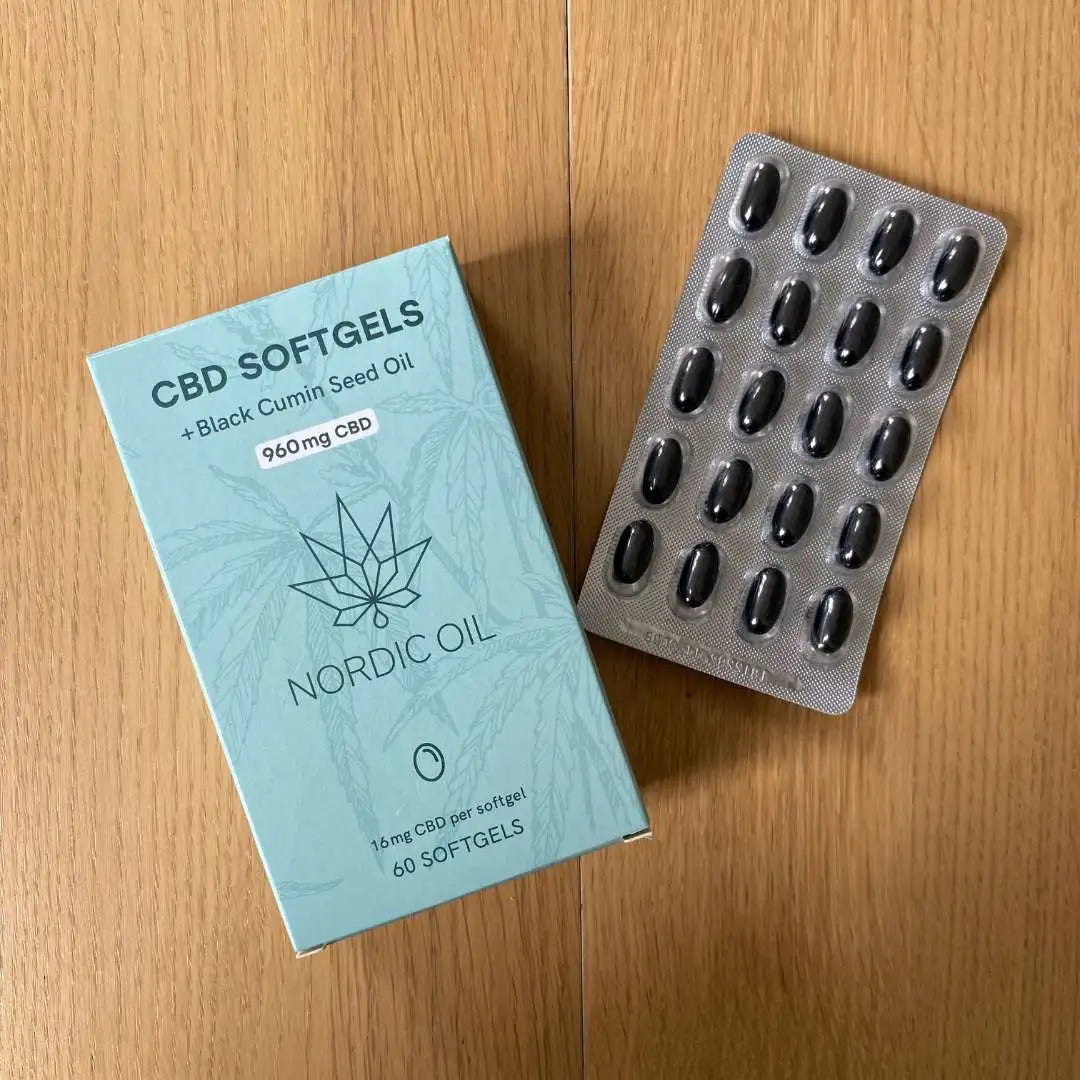 Lyseblå CBD-softgelboks ved siden af en blisterpakning med sorte kapsler.