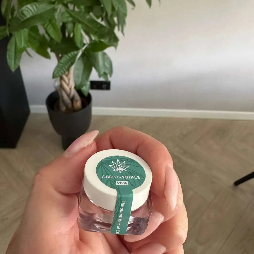 En hånd holder en lille glasbeholder med hvidt låg og grøn etiket, der læser CBD CRYSTALS 99%.