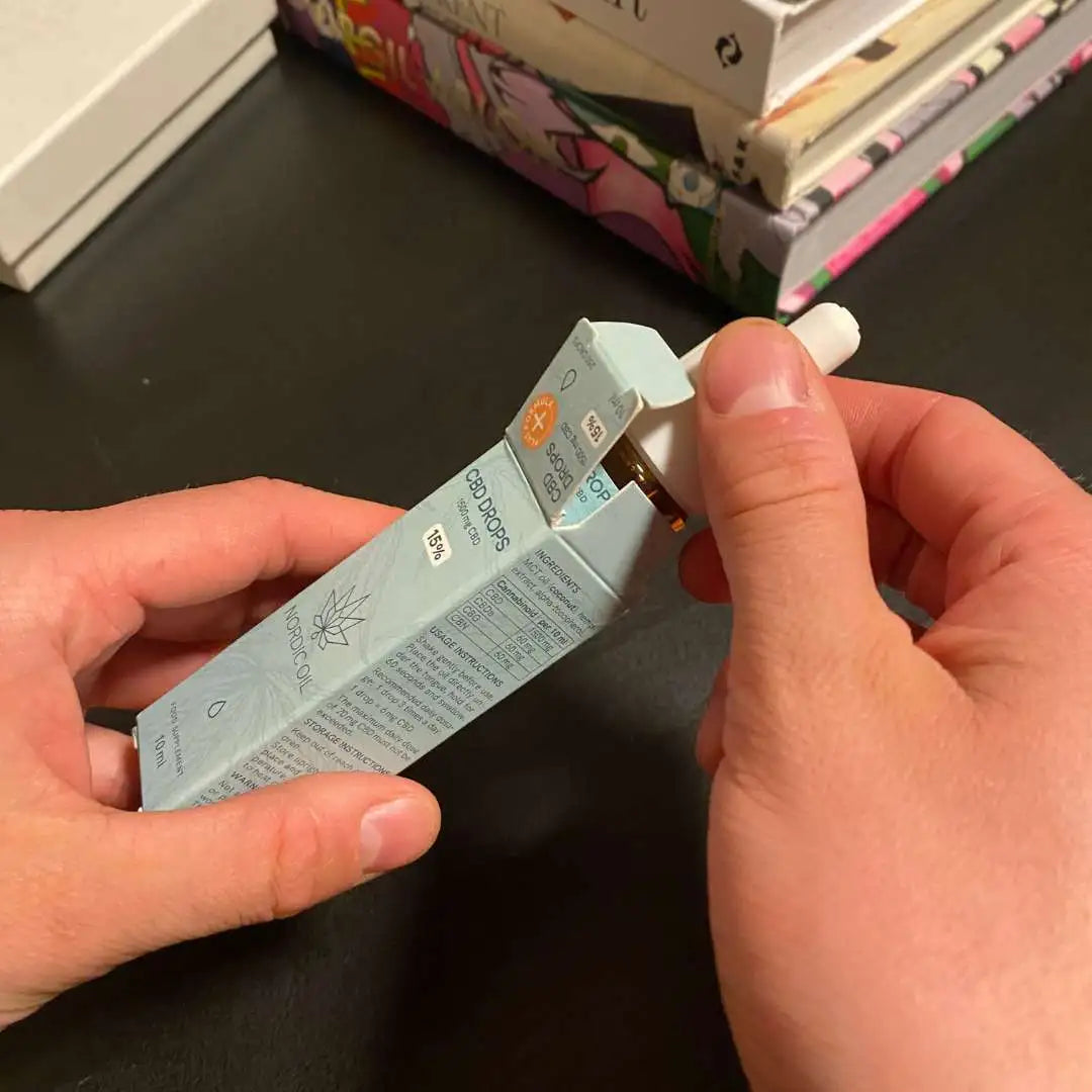 Nærbillede af hænder, der åbner en blå CBD-dråber-emballage med pipette.