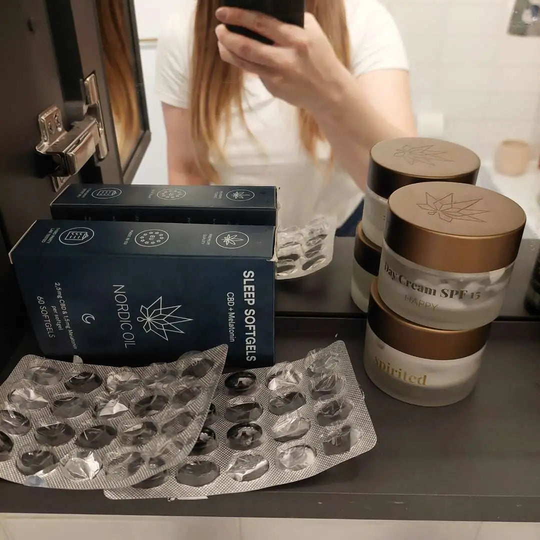 Person står ved spejlet i badeværelset og viser Nordic Oil Sleep Softgels-bokse samt to krukker Day Cream SPF15.