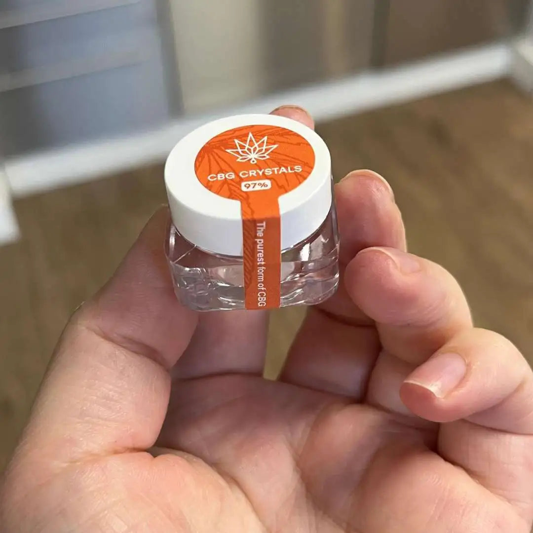 Lille glas med CBG-krystaller og hvidt låg med orange mærkat holdt mellem fingrene.