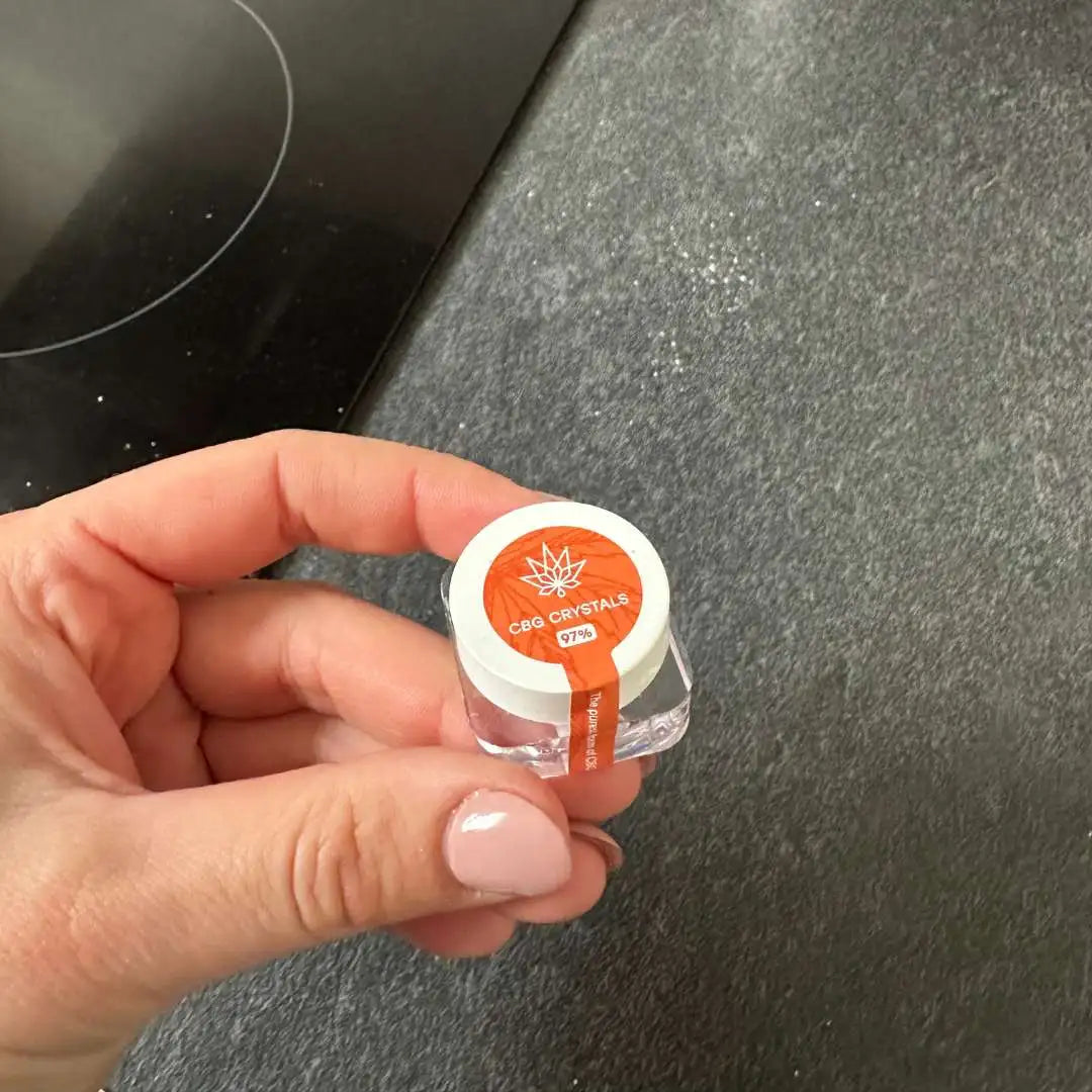 Hånd der holder en lille glasbeholder med CBD-kristaller og orange etiket