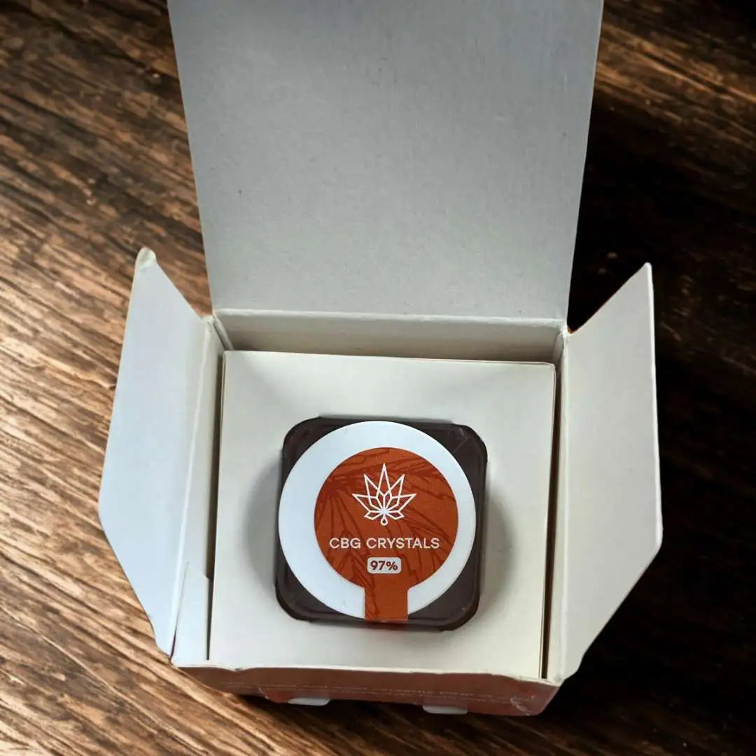 Åben æske indeholder lille sort beholder med orange etiket 'CBG CRYSTALS 97%'.