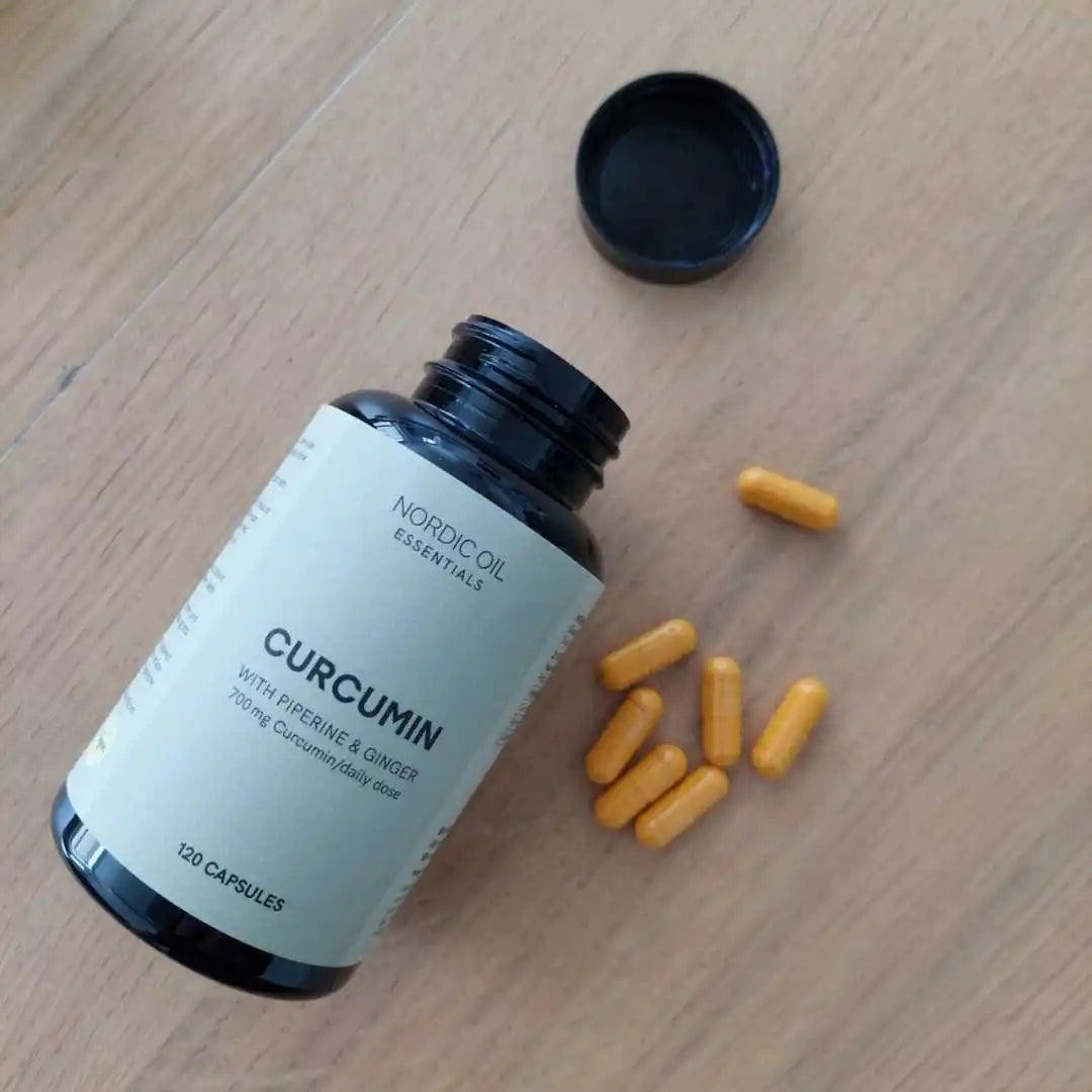 Nordic Oil Curcumin-kapsler i en mørk flaske med åbent låg liggende på et træbord sammen med gule kapsler.