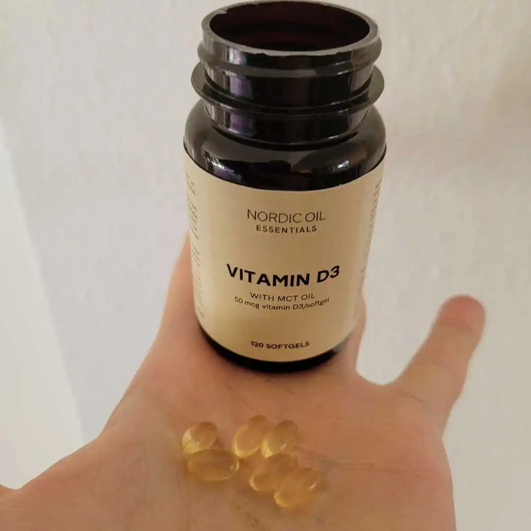 Hånd, der holder en flaske Nordic Oil Essentials Vitamin D3 med softgel-kapsler.