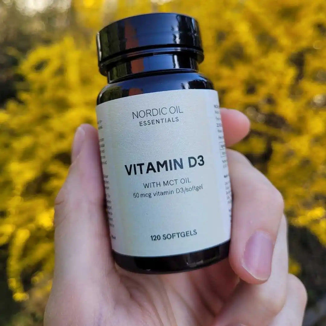Nærbillede af en Nordic Oil Essentials Vitamin D3-softgel-flaske, holdt i en hånd med gul baggrund.