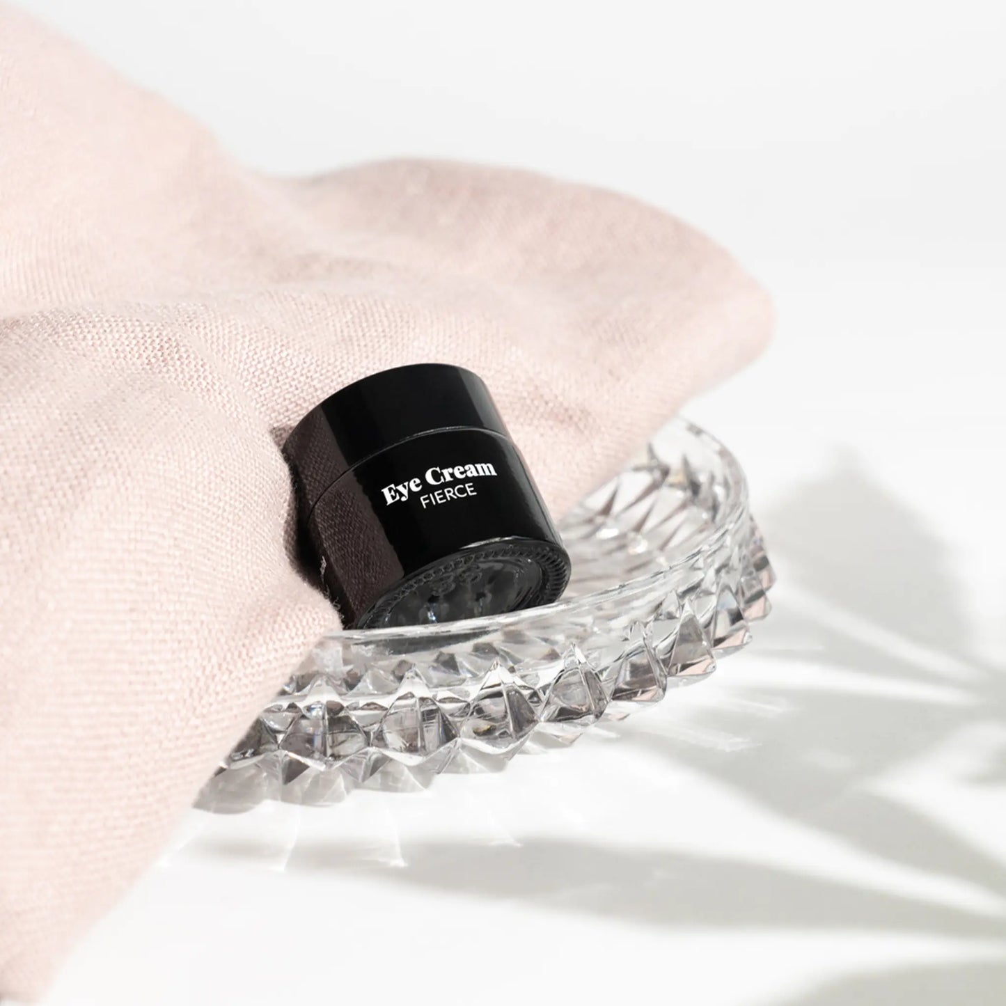 Sort øjencremekrukke mærket Eye Cream Fierce hviler på et facetteret krystalfad med lyserødt stof i baggrunden