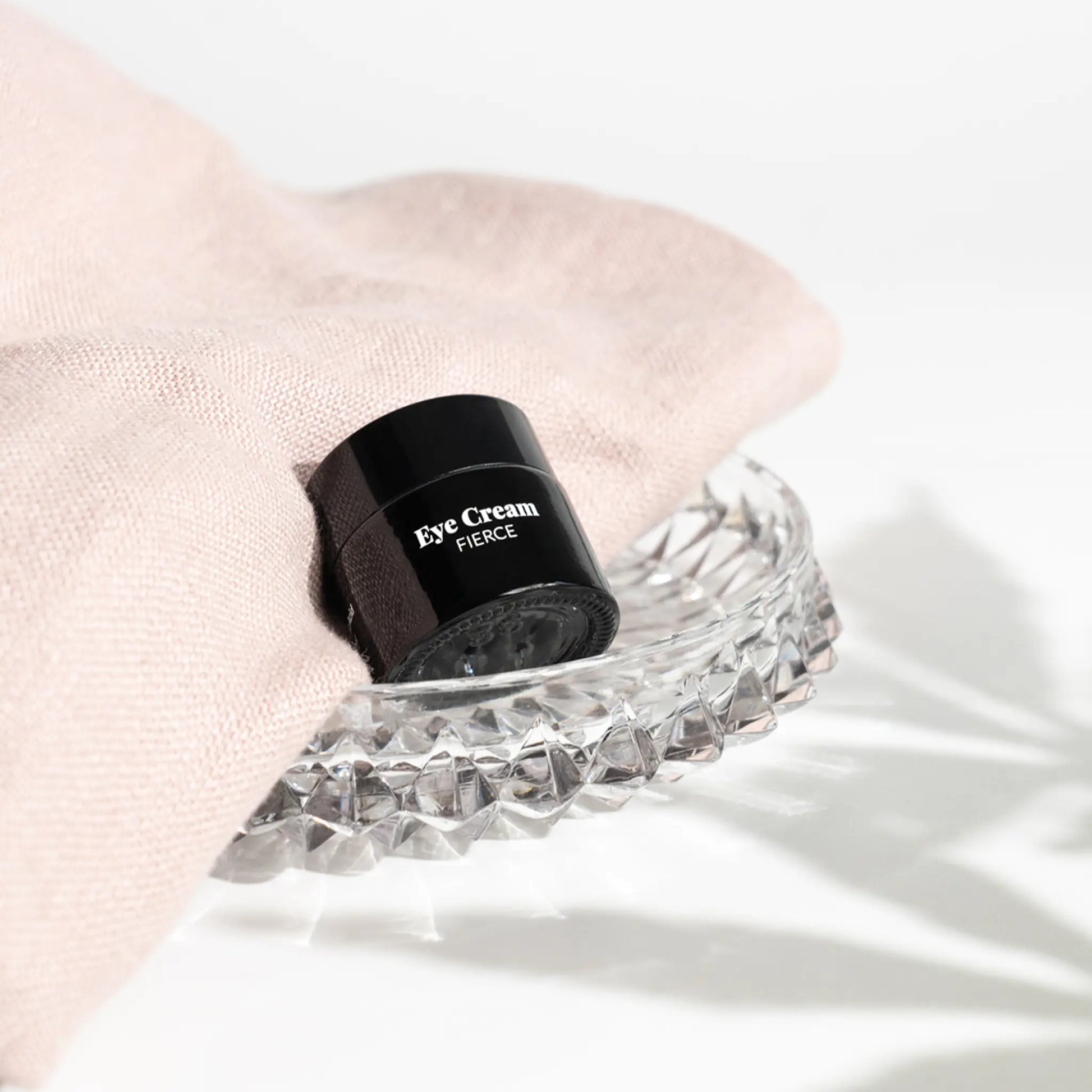 Sort øjencremekrukke mærket Eye Cream Fierce hviler på et facetteret krystalfad med lyserødt stof i baggrunden