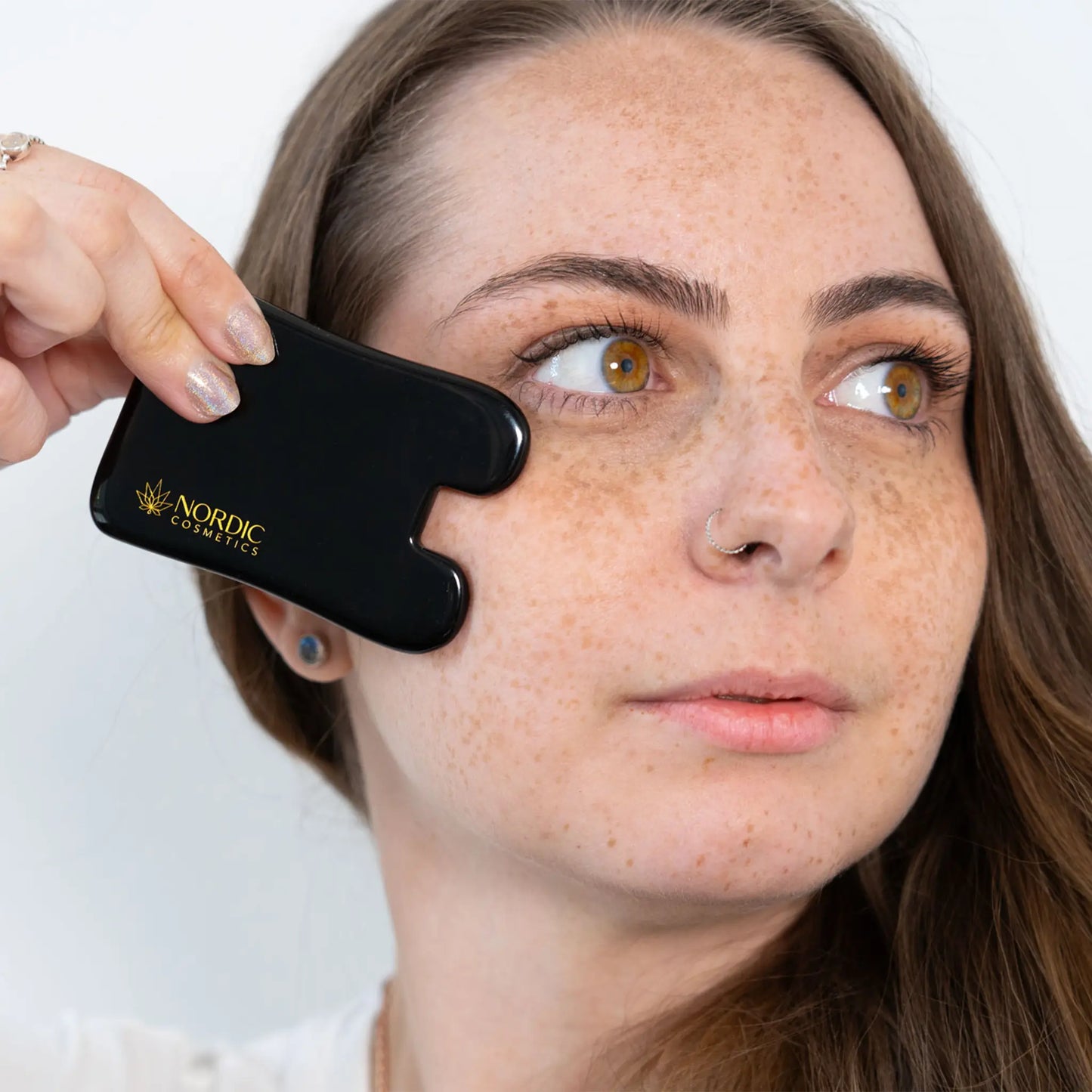 Kvinde holder et sort ansigtsmassageapparat med Nordic Cosmetics-logo tæt ved ansigtet.