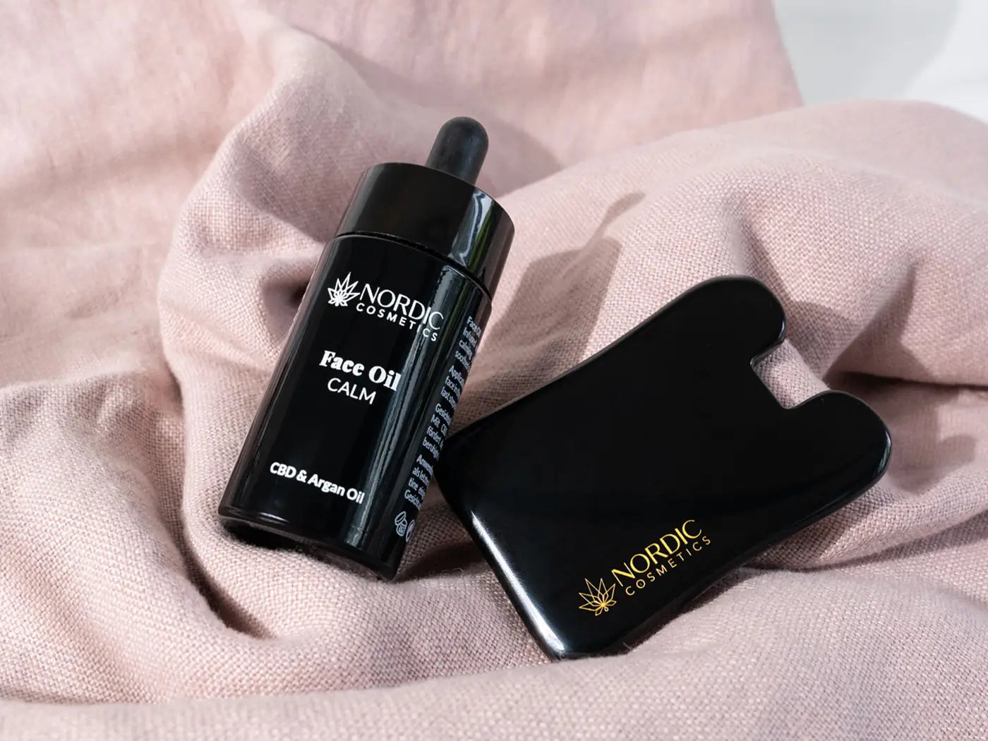 Nordic Cosmetics Face Oil Calm CBD og Argan Oil i sort flaske med dropper på lyserødt stof ved siden af sort tilbehør