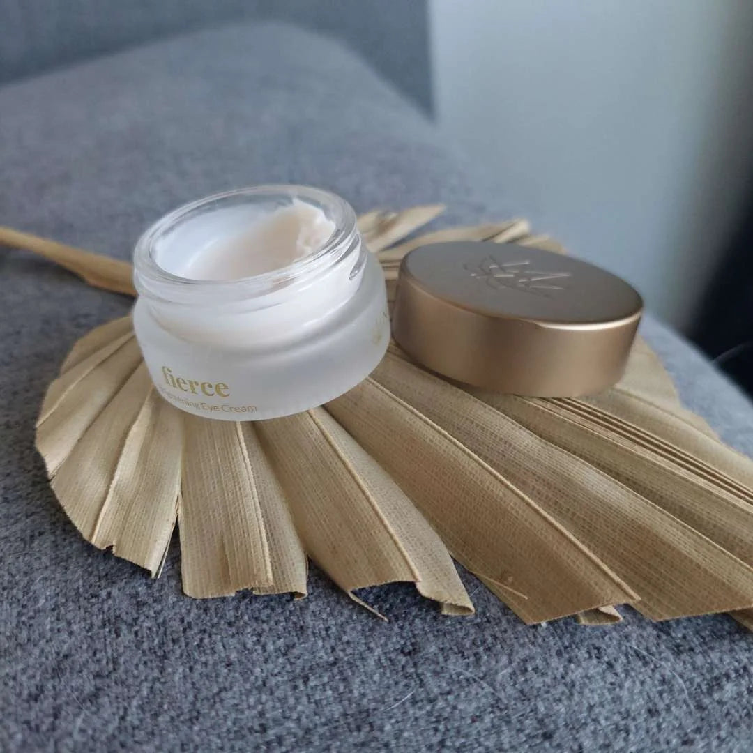 Lille åben glasbeholder med hvid øjencreme og gyldent låg ved siden af et tørret palmeblad.