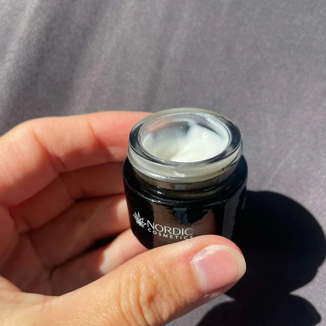 Lille sort krukke med hvid creme holdt i hånd, Nordic Cosmetics-logo synligt