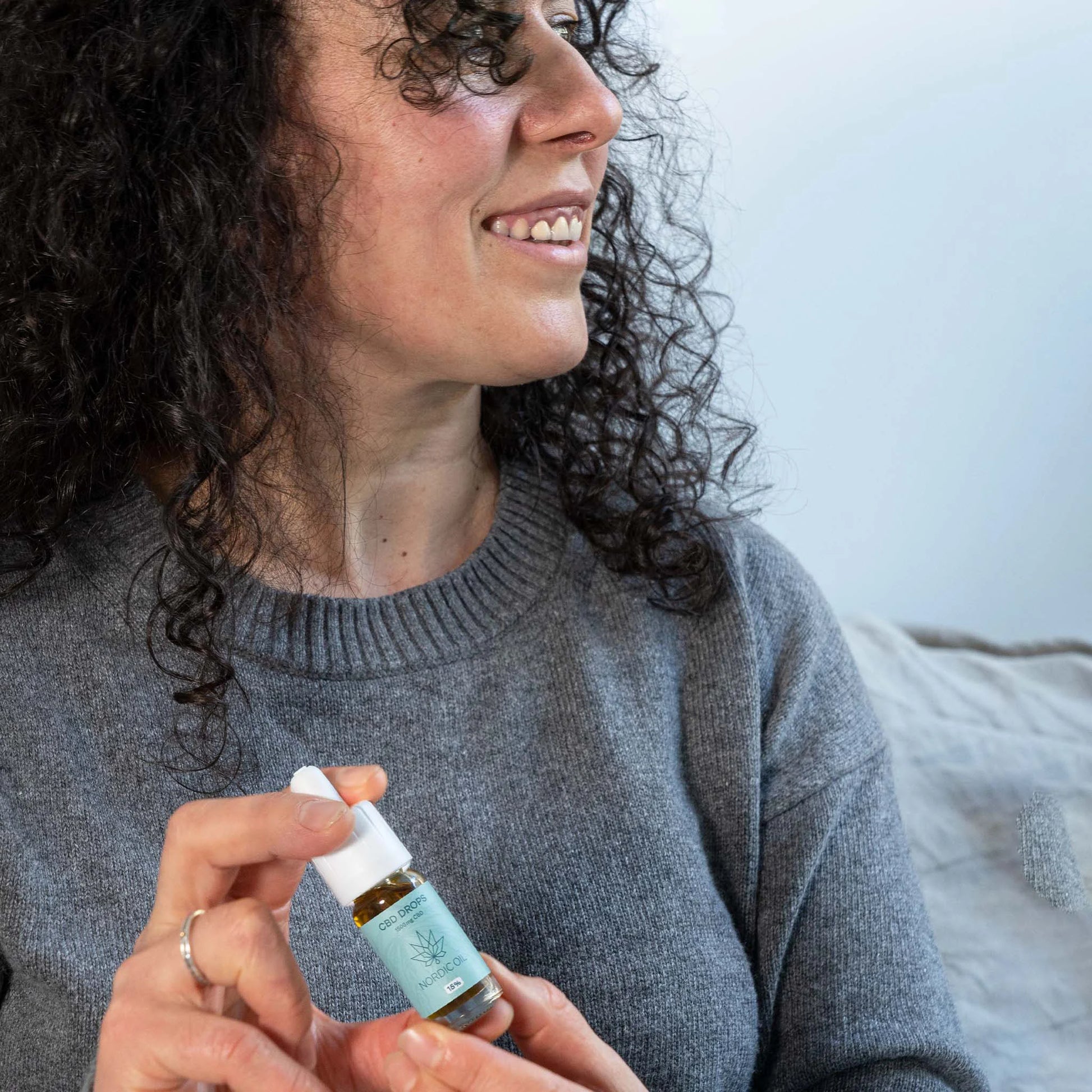 Kvinde med krøllet hår i grå trøje, der holder en lille flaske Nordic Oil CBD-olie med dropper.