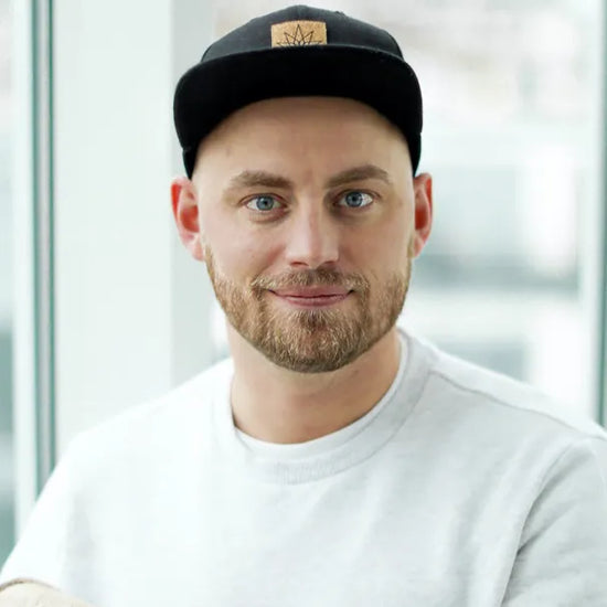Mand med skæg iført grå sweatshirt og sort kasket står ved vindue.