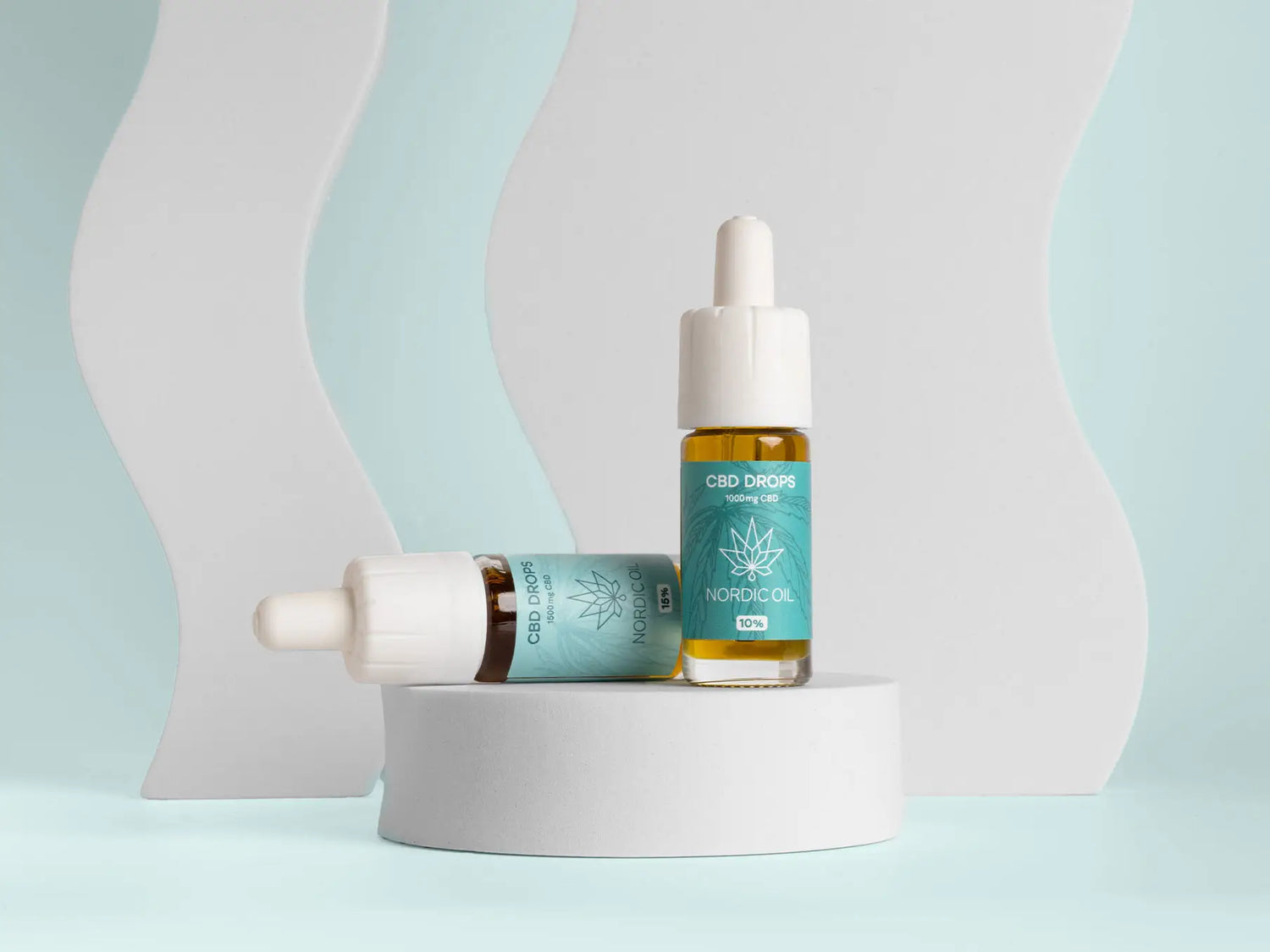Nordic Oil CBD-dråber – to glasflasker med blå etiketter på en hvid rund platform med pastelbaggrund