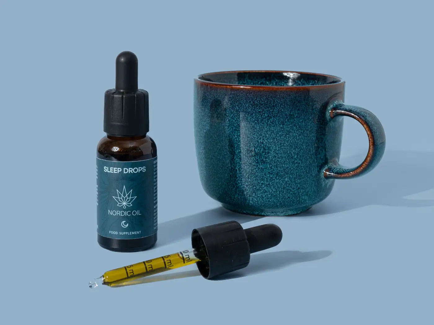 Nordic Oil Sleep Drops-flaske, en pipette og en blå keramikkrus på blå baggrund
