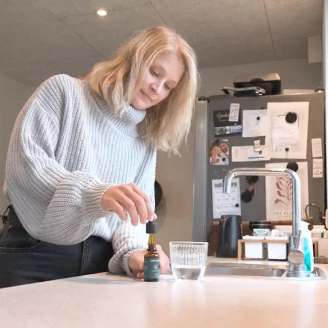 Kvinde i grå striktrøje hælder CBD-olie fra en dropper i et glas ved køkkenvasken.