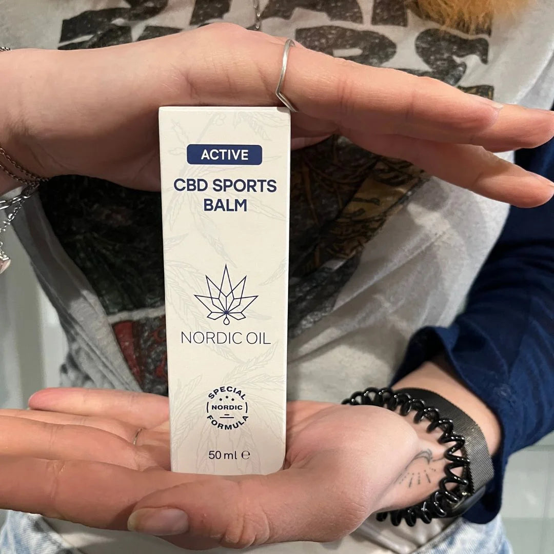 Nordic Oil CBD Sports Balm-æske i hvidt design med bladlogo, vist i hænderne (50 ml).