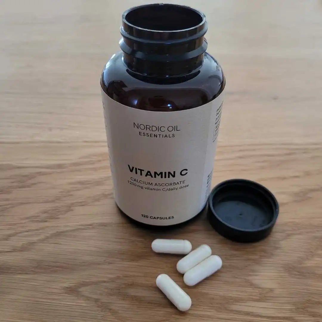 Nordic Oil Essentials Vitamin C kosttilskud i hvide kapsler ved siden af en åben låg på en brun glasflaske på et træbord